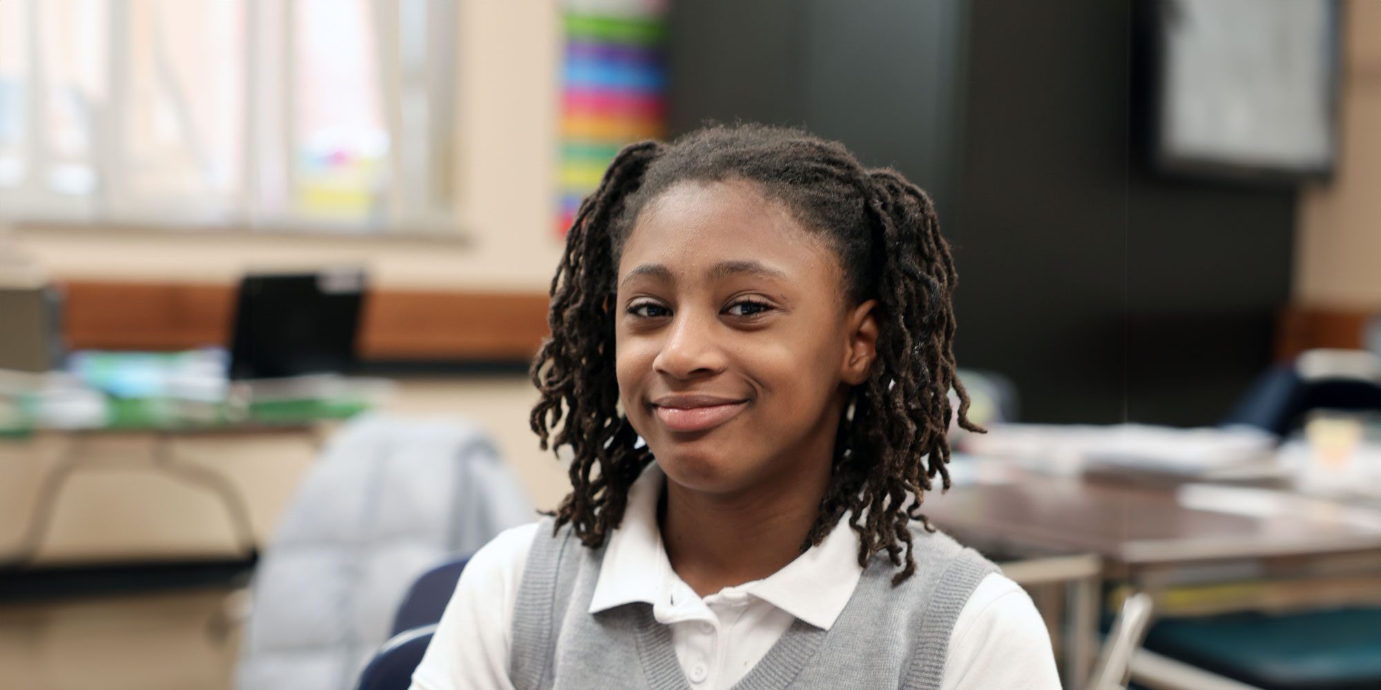 Smiling student in a gray uniform vest in a classroom.