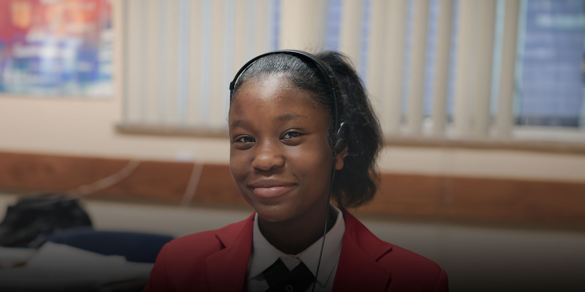 Smiling student in a red school blazer wearing headphones in a classroom.