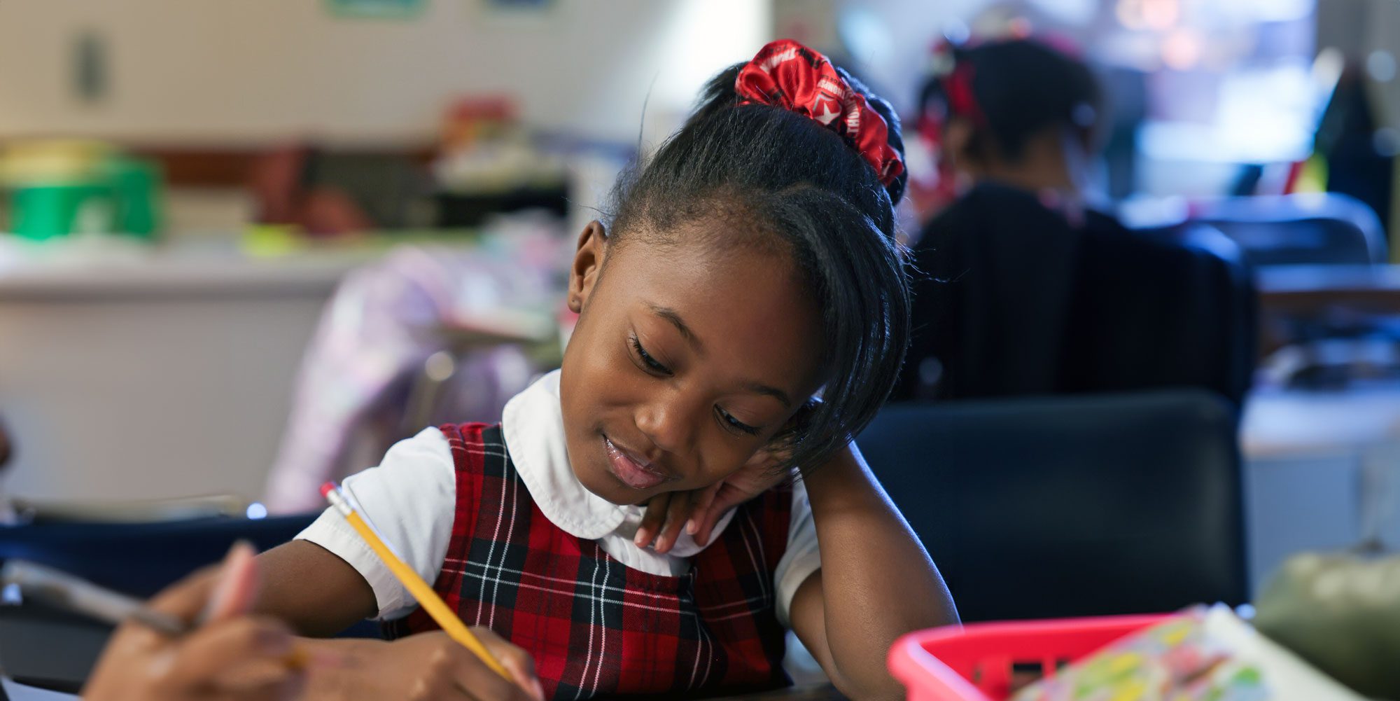 Student in a plaid uniform writing with a pencil at her desk.