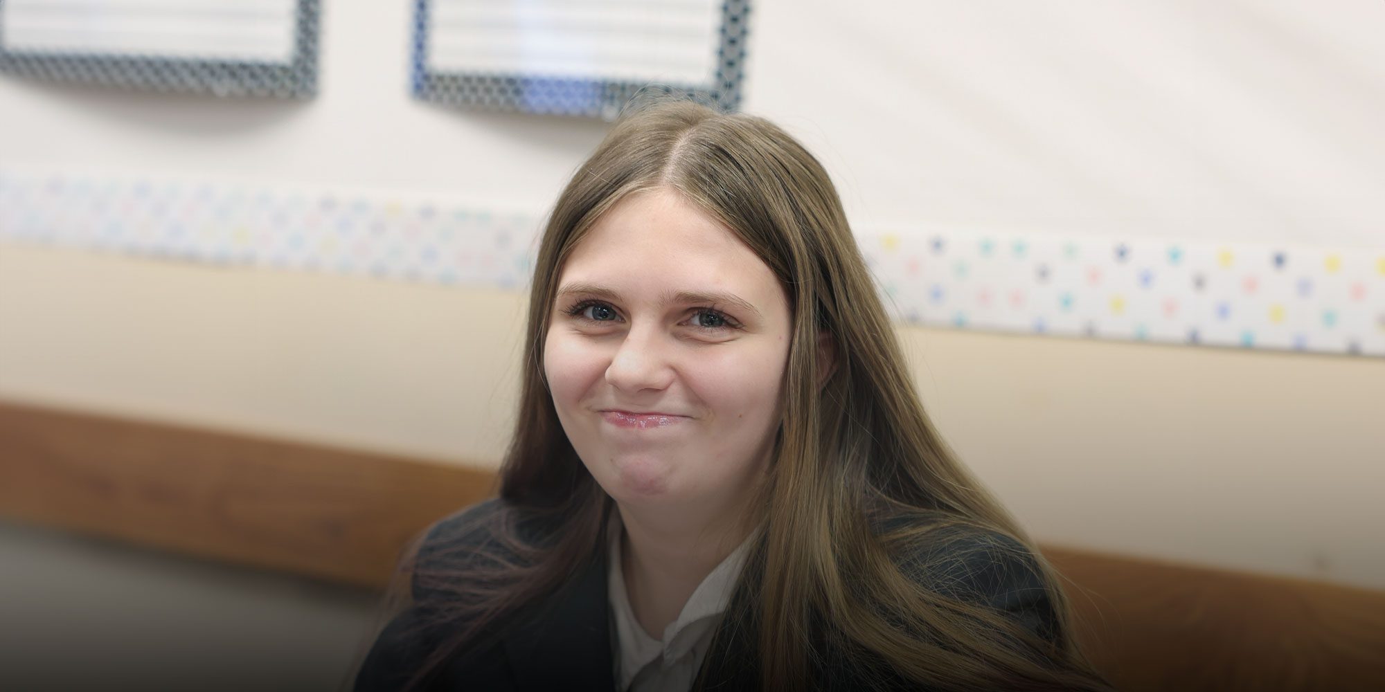 Smiling student in a school blazer in a classroom.